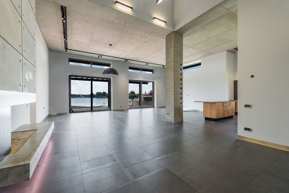 Stunning modern luxury home with large glass windows situated directly on the shore of a calm lake under a bright, sunny sky.